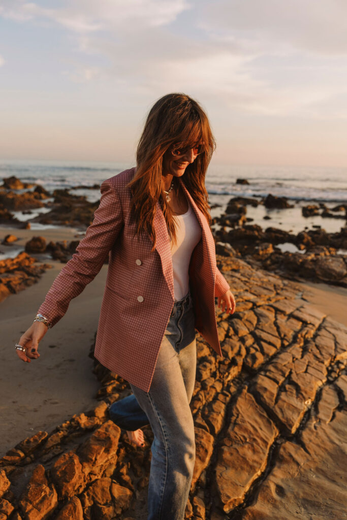 a women making more time for play on beach rocks in a peach blazer layed with a while top and blue jeans 