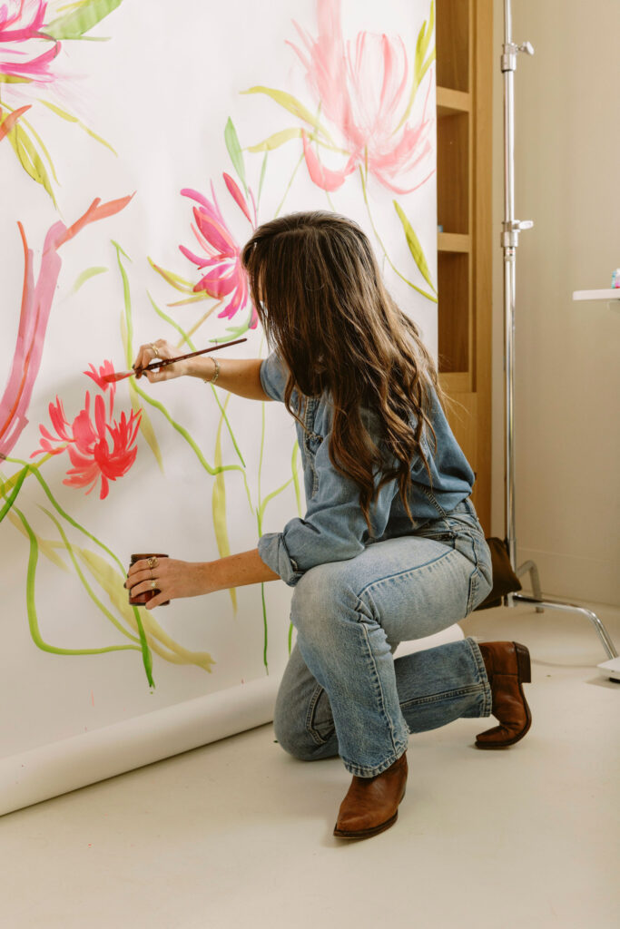 A woman with long brown hair, wearing a denim outfit and brown boots, kneels while painting large pink flowers on a white backdrop | cold press vs hot press watercolor paper