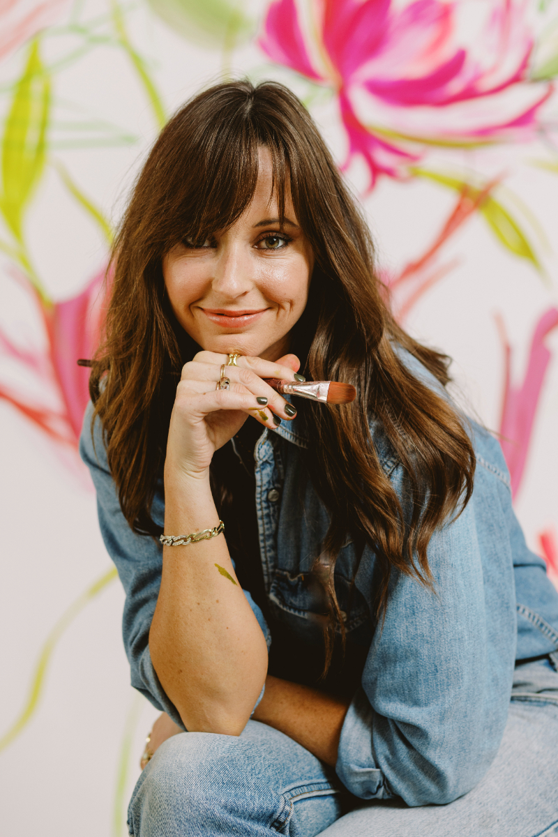 A smiling woman in denim holds a paintbrush, seated in front of a vibrant floral painting, radiating creative confidence.