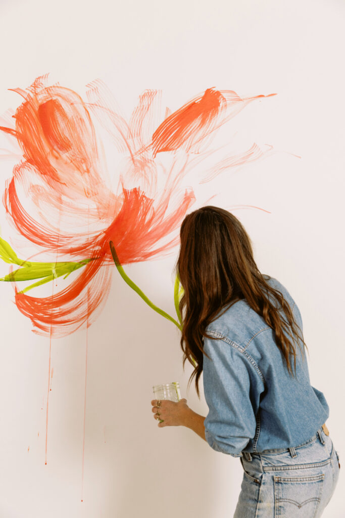 Woman in denim outfit paints a large red flower mural on a white wall using bold brushstrokes.