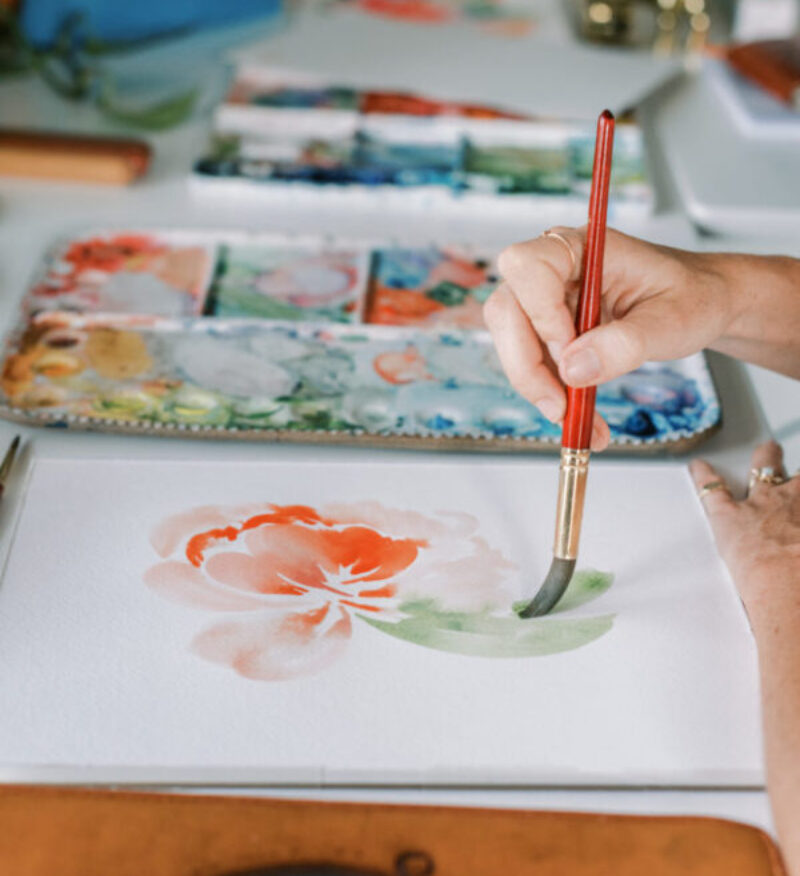 A hand uses a red paintbrush to create a soft, red-orange watercolor flower with green leaves, surrounded by painting supplies on a well-lit creative workspace.