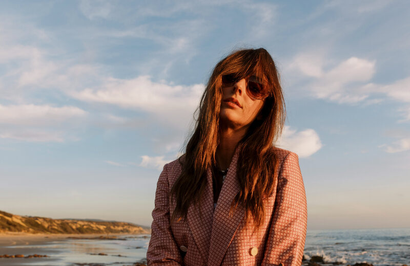 A woman wearing a pink patterned blazer and sunglasses stands on a rocky beach at sunset. She faces the camera with a relaxed pose, ocean and golden light glowing behind her.