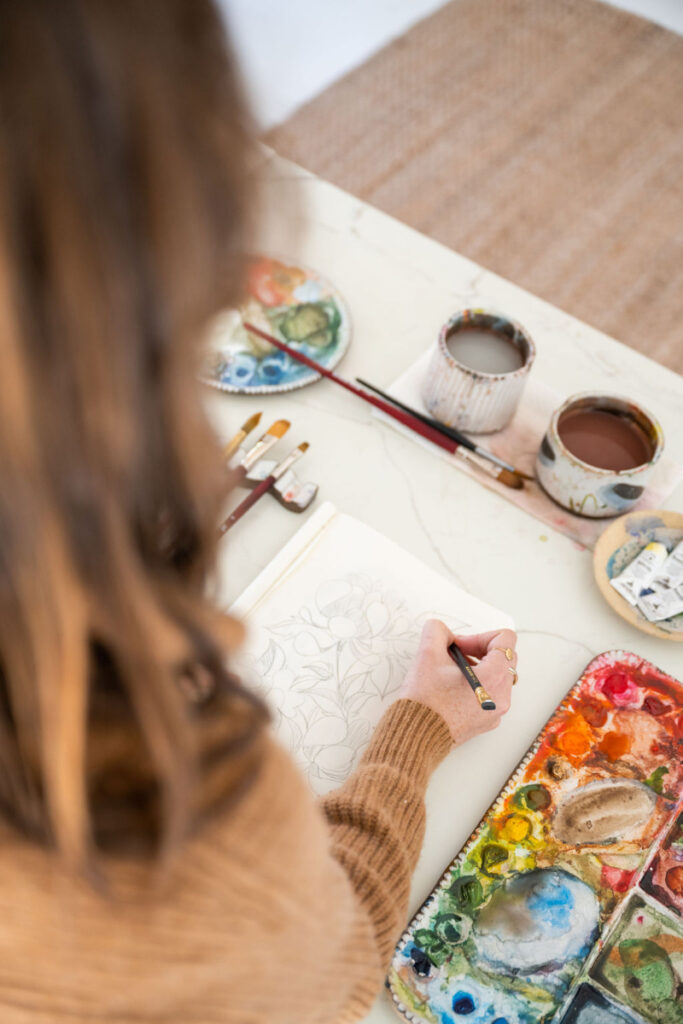 A top-down view of an artist sketching floral outlines in a sketchbook at a paint-covered table. Surrounding tools include brushes, watercolor palettes, and ceramic rinse cups.