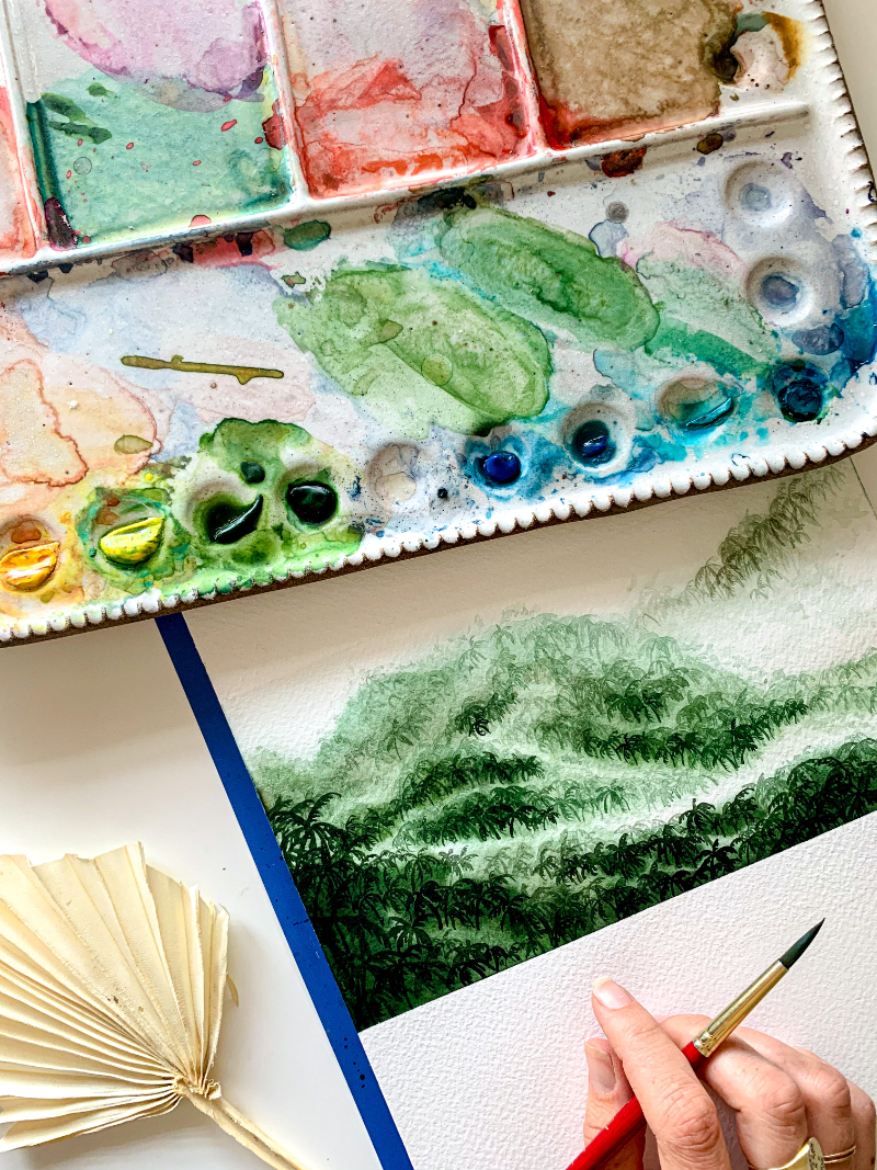 A watercolor palette filled with earthy tones and greens sits above a lush, layered green landscape painting in progress, with a red-handled brush held by a hand at the edge of the page.