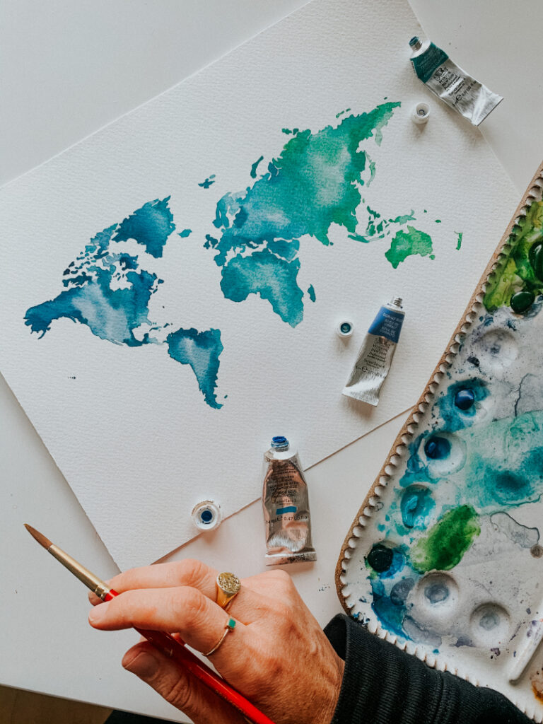 A hand holds a paintbrush beside a watercolor painting of a world map in blue and green tones, surrounded by paint tubes and a palette.