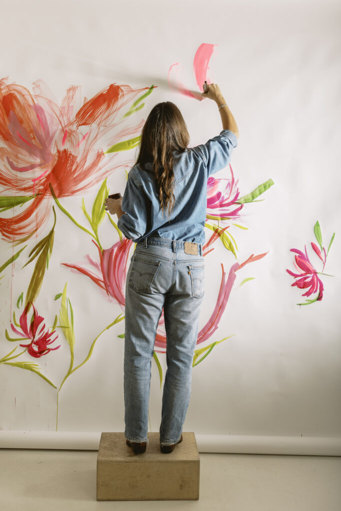 Artist in denim painting a large-scale floral mural with bold red and pink strokes.