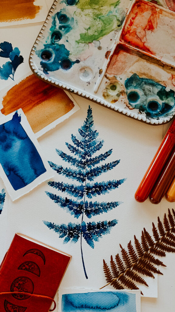 A detailed watercolor fall painting of a fern leaf in cool blue hues, surrounded by paint swatches, brushes, a palette, and pressed leaves, blending autumn nature with creative expression.