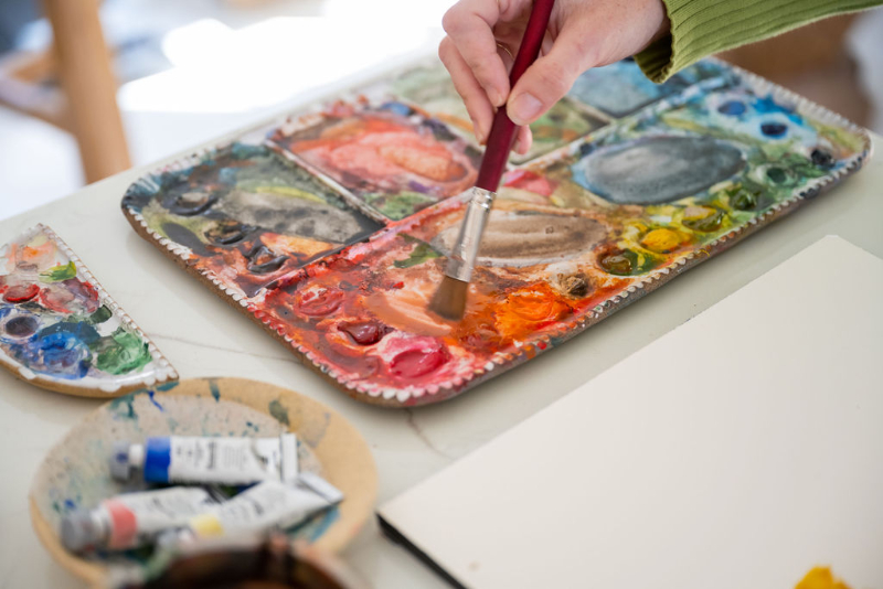 A hand mixing warm watercolor paints on a colorful palette beside paint tubes and paper, ready for a new scenery watercolor painting.