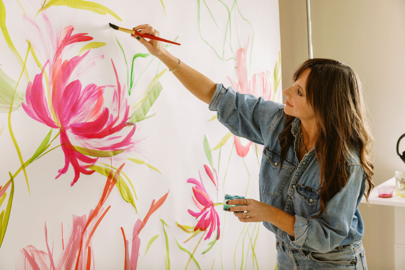 A woman painting large pink flowers on a white wall with broad watercolor brushstrokes while holding a small jar of paint.