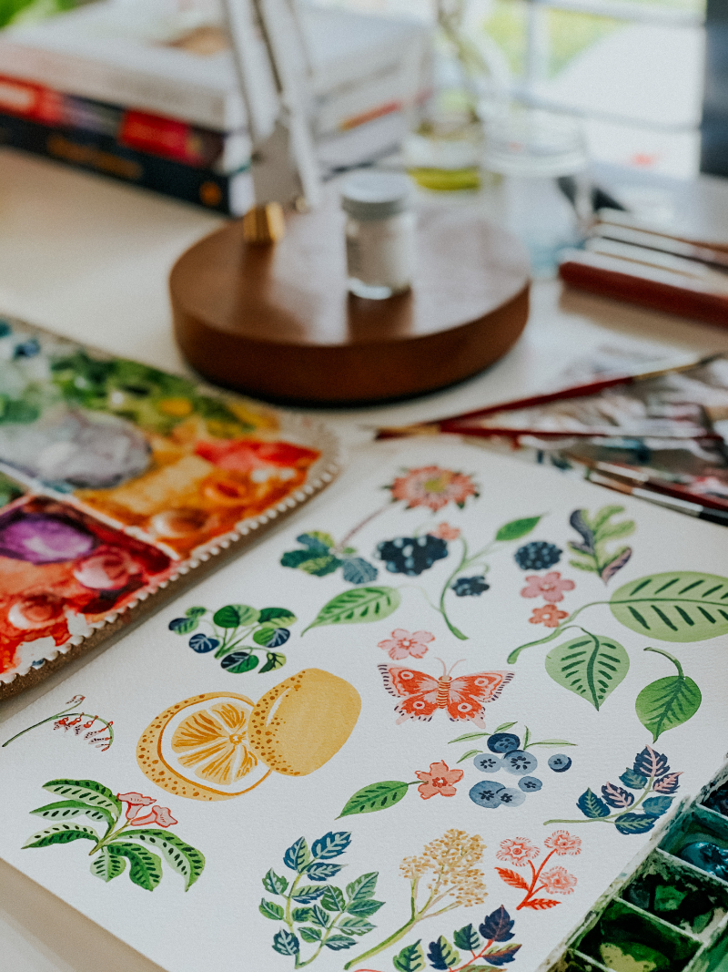 Close-up of a watercolor fruit and botanical painting with lemons, berries, butterflies, and leaves beside a colorful artist palette.