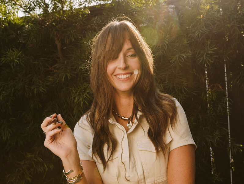 Woman smiling outdoors in soft sunlight, wearing a light-colored jumpsuit, holding a small object in her hand with greenery and bamboo plants behind her. | watercolor projects for valentine's day