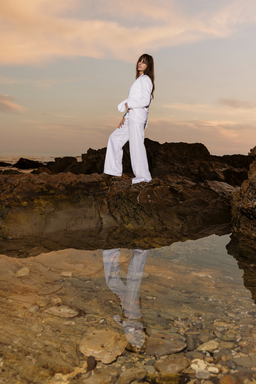 A woman in a white suit stands barefoot on coastal rocks at sunset, gazing confidently ahead. The tide pools below reflect her silhouette, creating a serene, introspective atmosphere against a soft pastel sky.
