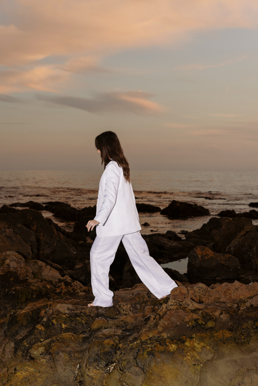 A woman in a white suit walks barefoot across rocky terrain near the ocean at dusk. The gentle pastel sky and reflective water create a quiet, contemplative mood as she moves through the landscape.