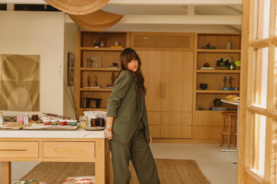 A woman in a dark green suit stands inside a warm, modern art studio filled with brushes, paints, and watercolor pieces. The setting feels grounded, expressive, and reflective of an artist reconnecting with her practice after creative burnout.