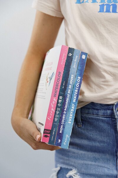 A person wearing a casual T-shirt and jeans holds a stack of watercolor books, including “Everyday Watercolor,” “Everyday Watercolor Workbook,” and international editions. The image conveys learning, creativity, and building watercolor skills through guided instruction.