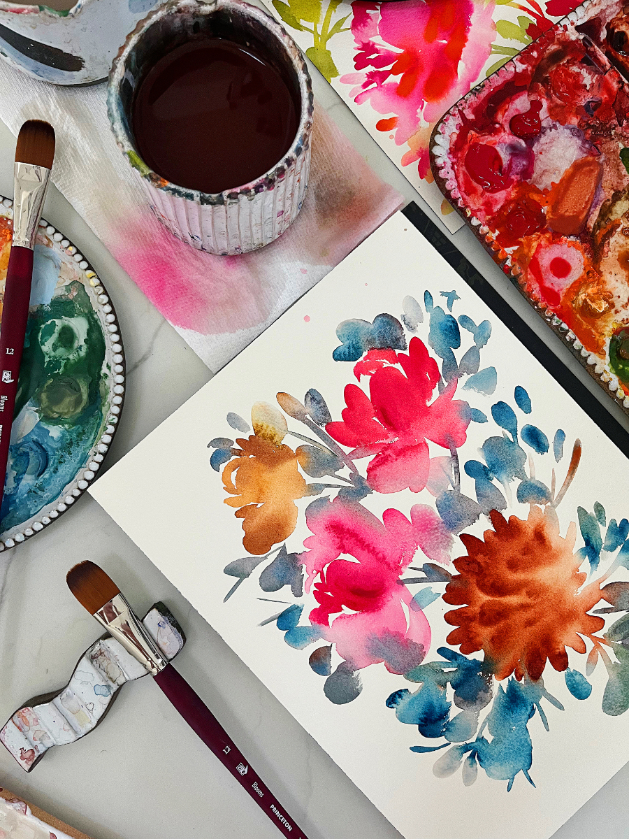A studio table filled with watercolor supplies, brushes, and floral artwork in progress. Bold pinks, oranges, and blues blend together on the page and palette, showing the luminous layering and fluid transparency that comes from painting watercolor light to dark.