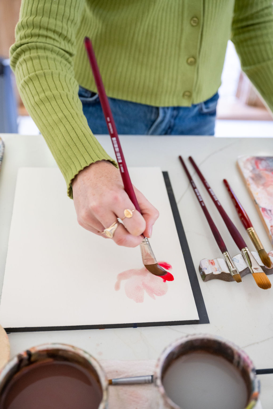 A person in a green sweater uses a large watercolor brush to apply soft red paint onto blank watercolor paper. Multiple clean brushes and cups of water sit nearby, capturing the first gentle wash typical when painting watercolor light to dark.