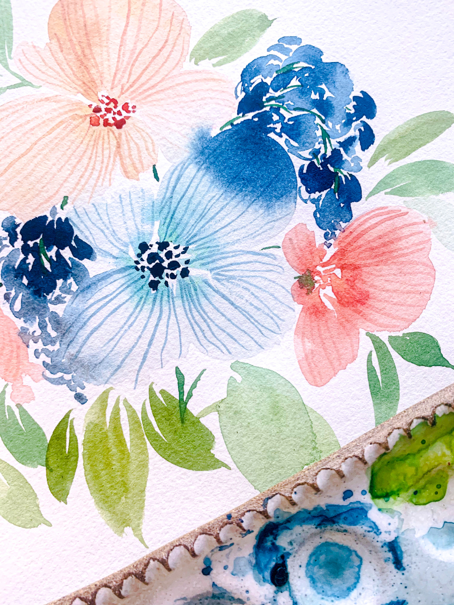 A close-up of a finished floral watercolor painting featuring blue, coral, peach, and green petals and leaves. The delicate layers show value shifts and transparency achieved through painting watercolor light to dark, with a ceramic mixing palette resting at the edge of the frame.
