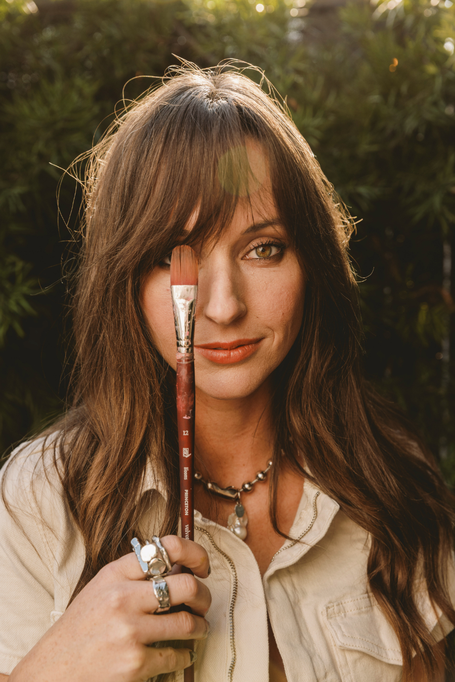 A close-up portrait of a woman holding a large watercolor brush in front of one eye while standing outdoors. Her expression is calm and confident, with warm natural light illuminating her face, suggesting a personal connection to creativity and her watercolor supplies.