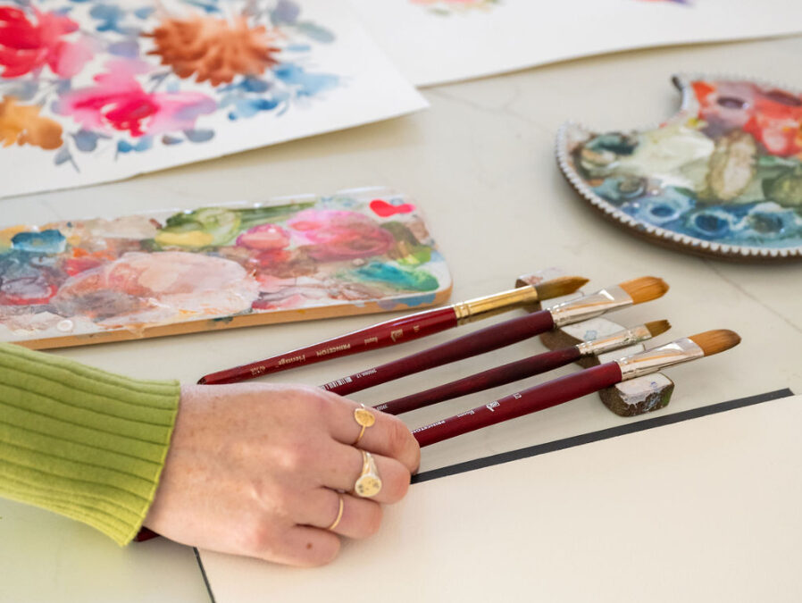 A hand reaches for a set of red-handled watercolor brushes beside a blank sheet of paper. Surrounding the workspace are colorful floral paintings, paint palettes, and scattered watercolor supplies that highlight the process and tools behind creating expressive botanicals.