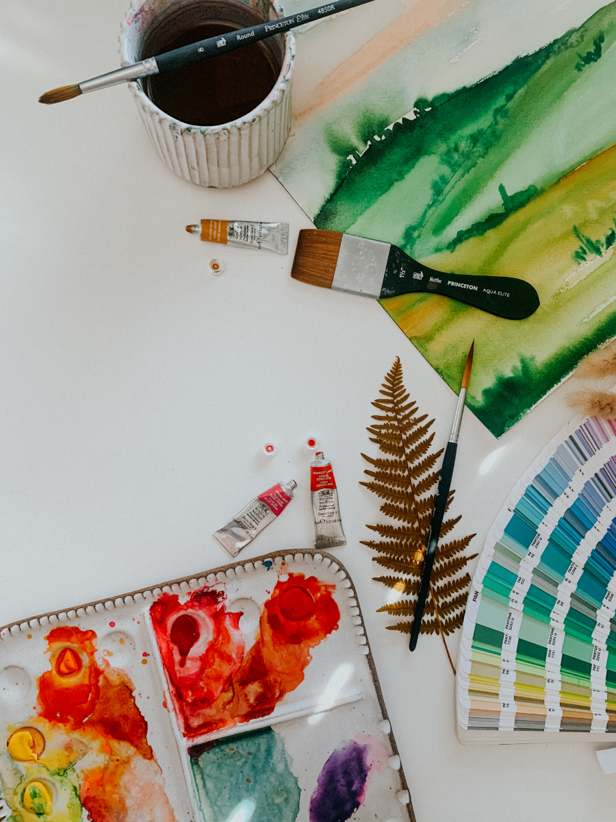 Overhead view of a watercolor workspace with a palette of vivid paints, brushes, and tubes scattered around. A green landscape wash dries beside a color swatch fan and a pressed fern, capturing an experimental moment in oil painting vs watercolor techniques.
