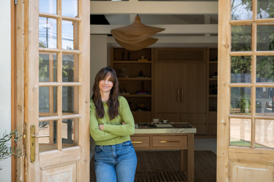 A woman in a green sweater stands in a sunlit doorway of a warm, modern studio with wooden details. She smiles with arms crossed, framed by open glass doors, offering a welcoming glimpse into a calm, creative workspace.