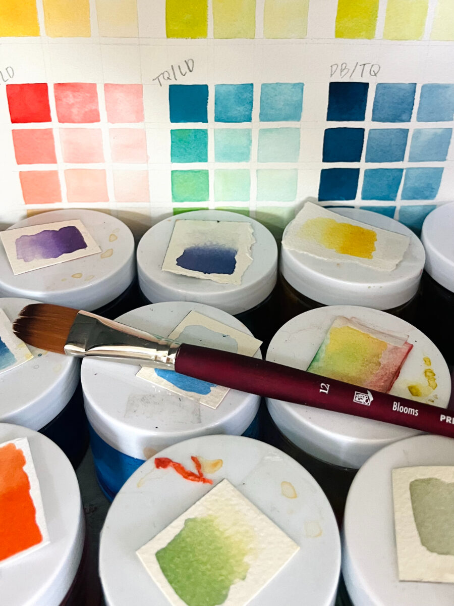 A detailed color study display with neatly arranged watercolor swatches in gradients of greens, blues, and yellows. Small sample papers rest on jars below, alongside a brush, showcasing careful mixing, testing, and organized artistic exploration.