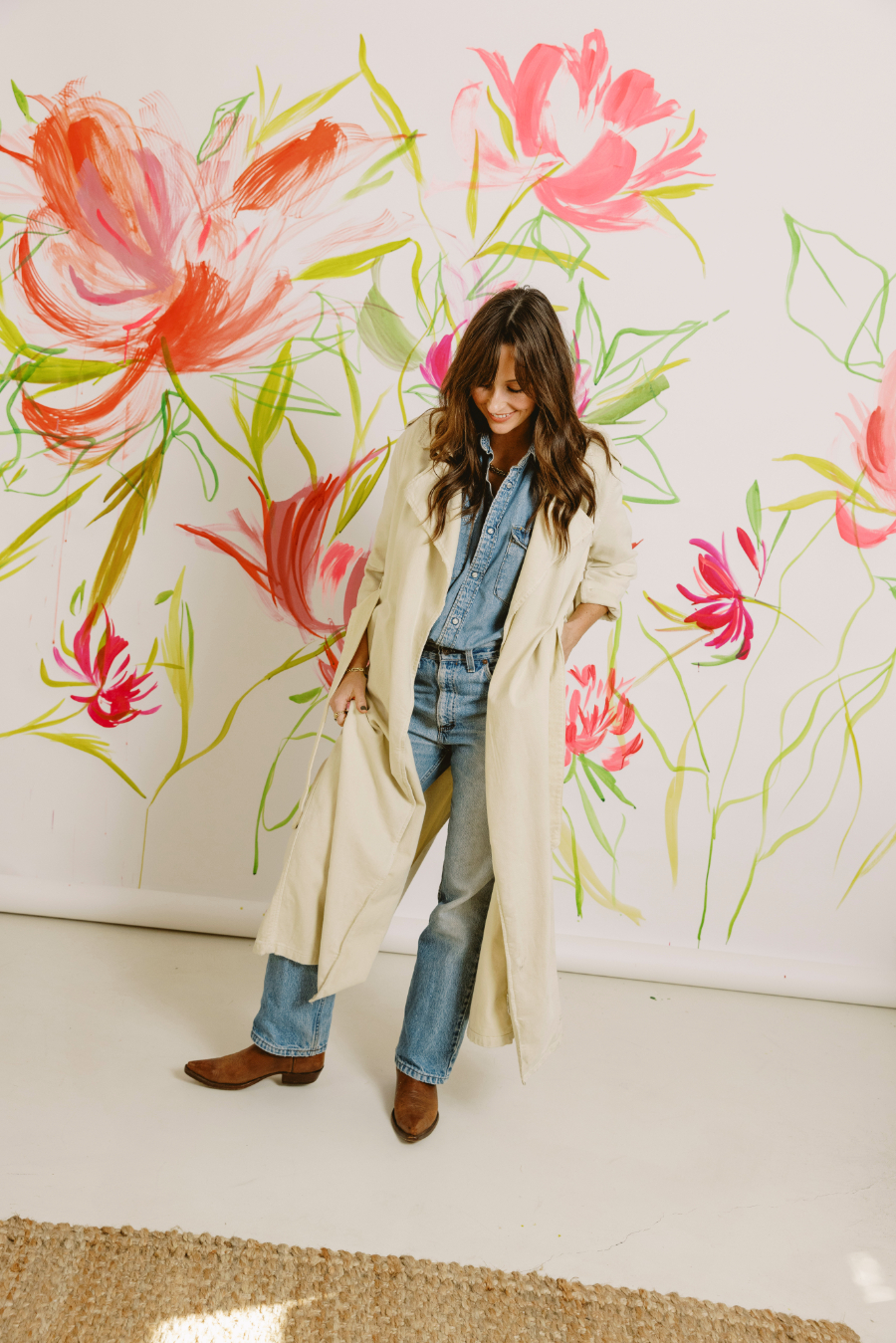 A woman stands in front of a large wall painted with expressive, oversized floral strokes in pinks and greens. Wearing a long coat and denim, she looks down with a relaxed smile, blending fashion, creativity, and bold artistic expression.