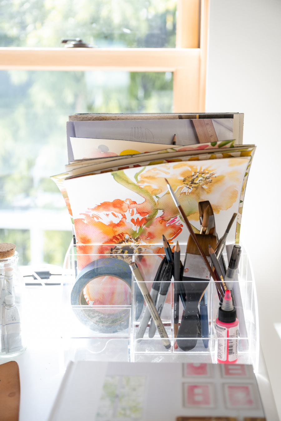 Sunlit window workspace with a clear acrylic organizer holding watercolor paintings, brushes, pens, and tape. Floral artwork in warm orange tones is visible among stacked papers, creating a calm, creative studio atmosphere filled with natural light.