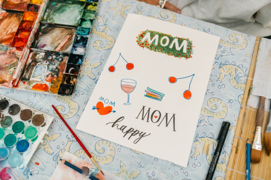 Hand-painted watercolor sheet featuring colorful illustrations and lettering reading “Mom,” surrounded by art supplies. The composition highlights a playful, heartfelt design process for handmade mother’s day cards on a patterned surface.