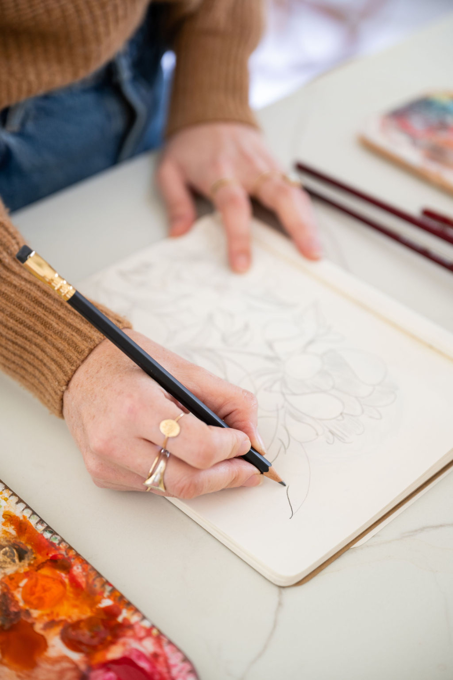 Close-up of an artist sketching floral outlines in a notebook beside a watercolor palette. This planning stage helps establish composition and placement, guiding where the focal point in art will sit before adding color and detail.