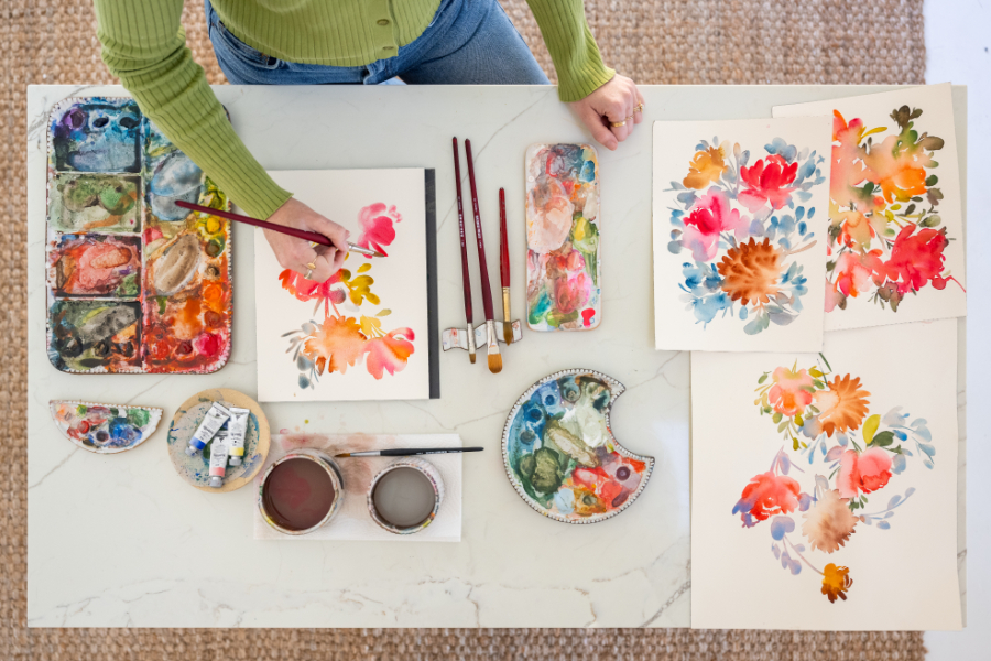 Overhead view of an artist painting loose watercolor florals surrounded by palettes and brushes. Multiple compositions show how varying size, color, and placement can create a strong focal point in art while keeping the overall design balanced and expressive.