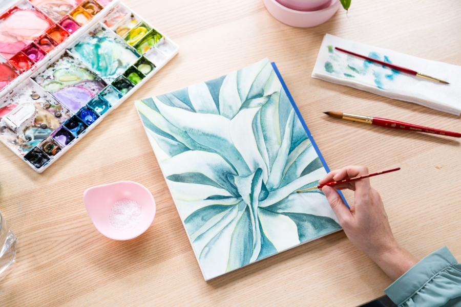Artist painting a soft green botanical composition with layered watercolor washes. Subtle value shifts and crisp edges draw attention to the center, illustrating how a focal point in art can be created through contrast, brush control, and intentional detail.