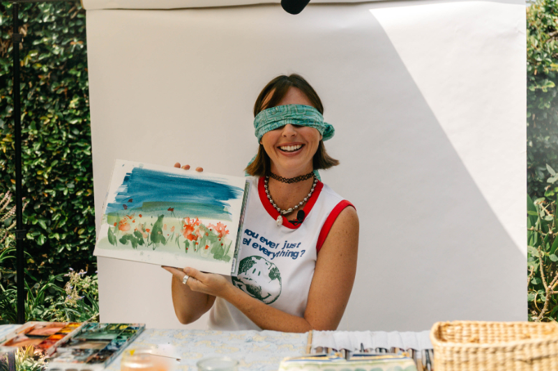 A smiling artist wearing a blindfold holds up a watercolor landscape of flowers under a deep blue sky. Surrounded by paints and brushes outdoors, she playfully explores creativity and intuition—an imaginative approach to painting a night scene without overthinking technique.