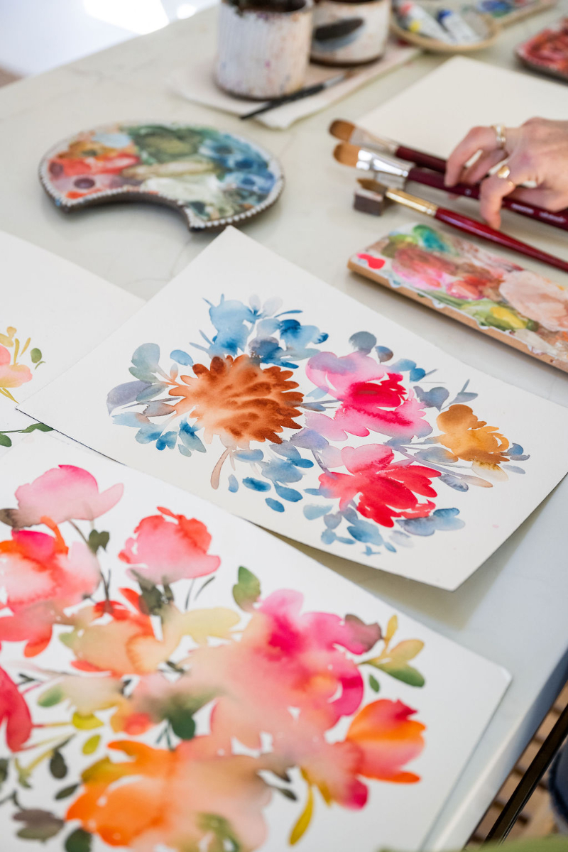 Several watercolor floral paintings lie on a studio table beside brushes and palettes. The compositions feature vivid pinks, blues, and oranges, showcasing painting with values through layered tones and contrast that bring each bloom to life.