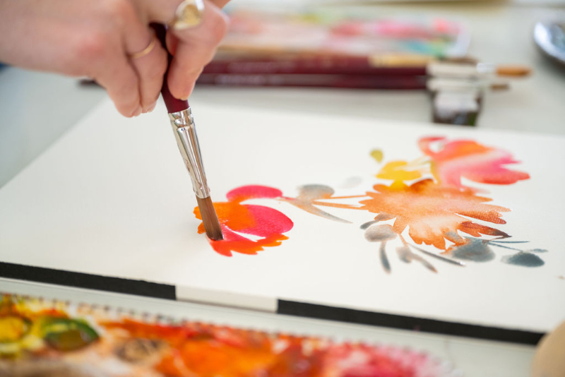 A hand holds a paintbrush, blending warm reds, oranges, and grays into soft floral shapes on watercolor paper. The image captures the subtle tonal shifts that define painting with values—balancing light and dark to create depth and vibrancy.
