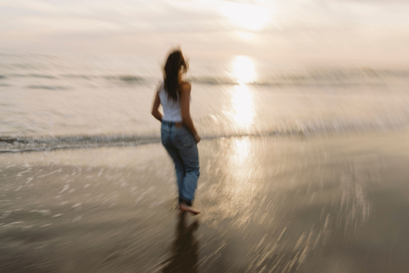 A woman walks barefoot along the shoreline at sunset, her reflection shimmering on wet sand. The soft motion blur and golden light evoke the tranquil atmosphere of watercolor beach scenes and coastal inspiration.