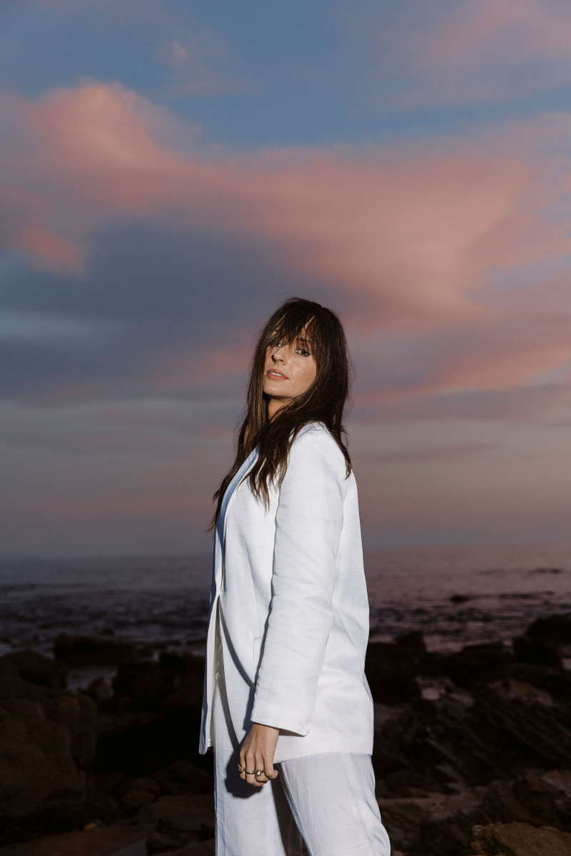A woman in a white linen suit stands on a rocky shore at dusk, gazing toward the camera as soft pink and blue clouds drift over the ocean.