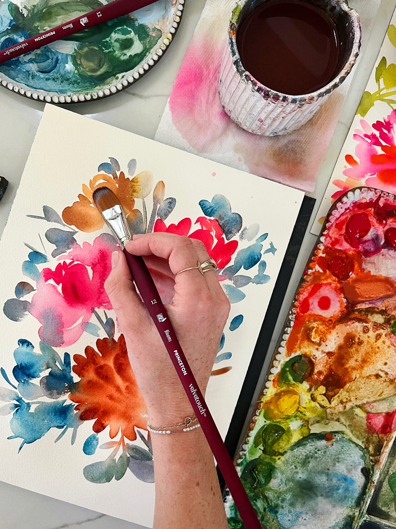 Hand painting a vibrant floral composition with watercolor brushes, palette, and supplies spread across a workspace.