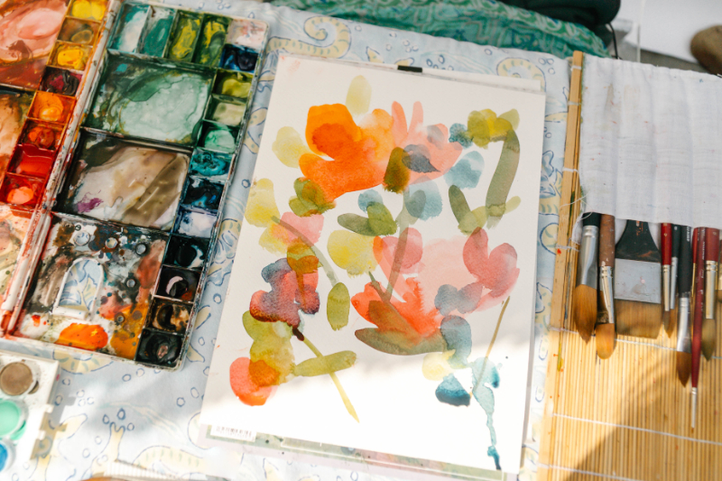 Loose watercolor floral painting created during a blindfold challenge, surrounded by watercolor palettes, brushes, and art supplies.