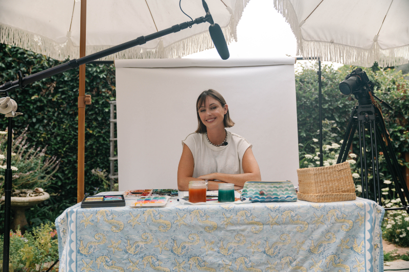 Artist filming a watercolor tutorial at an outdoor table with paints and supplies, perfect inspiration for a watercolor paint night.