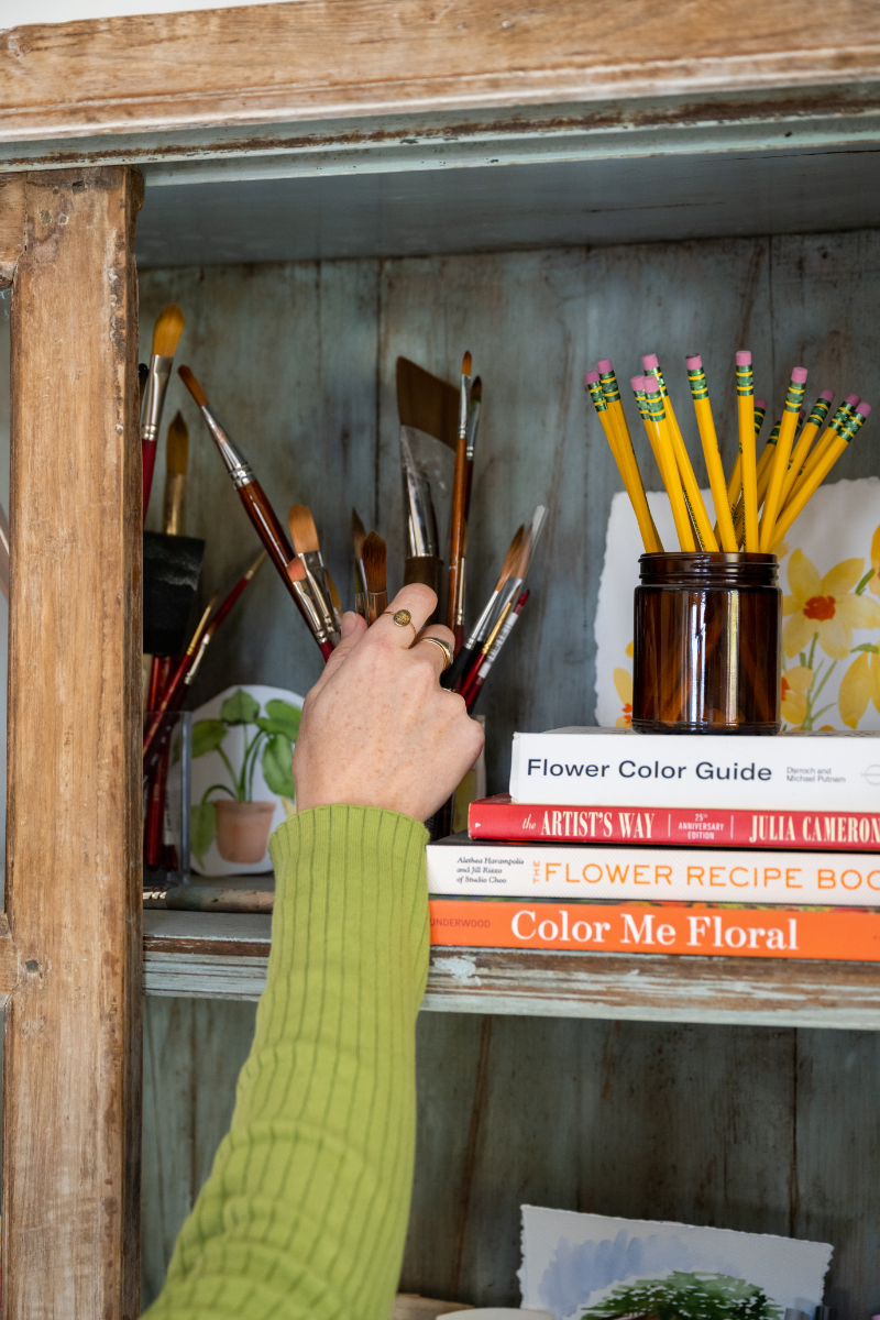 Collection of watercolor brushes, pencils, and art books arranged on a shelf with supplies ready for a watercolor paint night.