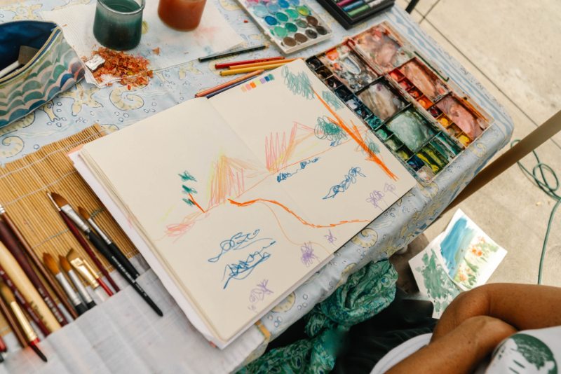 An open sketchbook filled with colorful hand-drawn illustrations sits beside watercolor paints and brushes, highlighting the creative process behind ai art vs digital art.