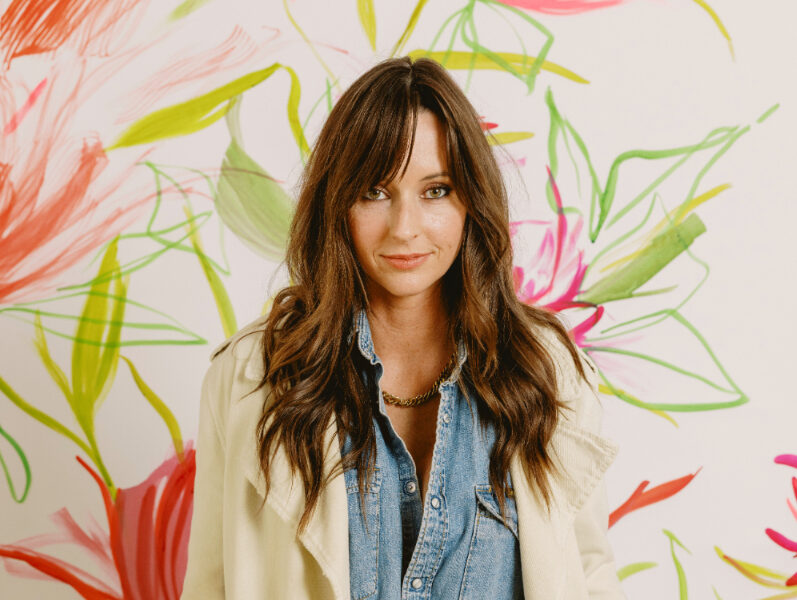 Jenna Rainey stands in front of a vibrant floral mural wearing a denim outfit and cream coat, inspiring artists to develop their own creative style through a beginner painting tutorial.