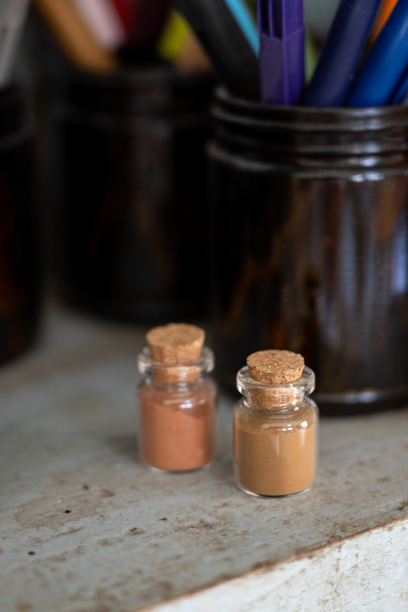 Two small glass bottles filled with warm brown and terracotta pigments sit on an artist’s studio shelf beside containers of colorful pens, highlighting pigment quality and materials when comparing cheap vs professional watercolor.