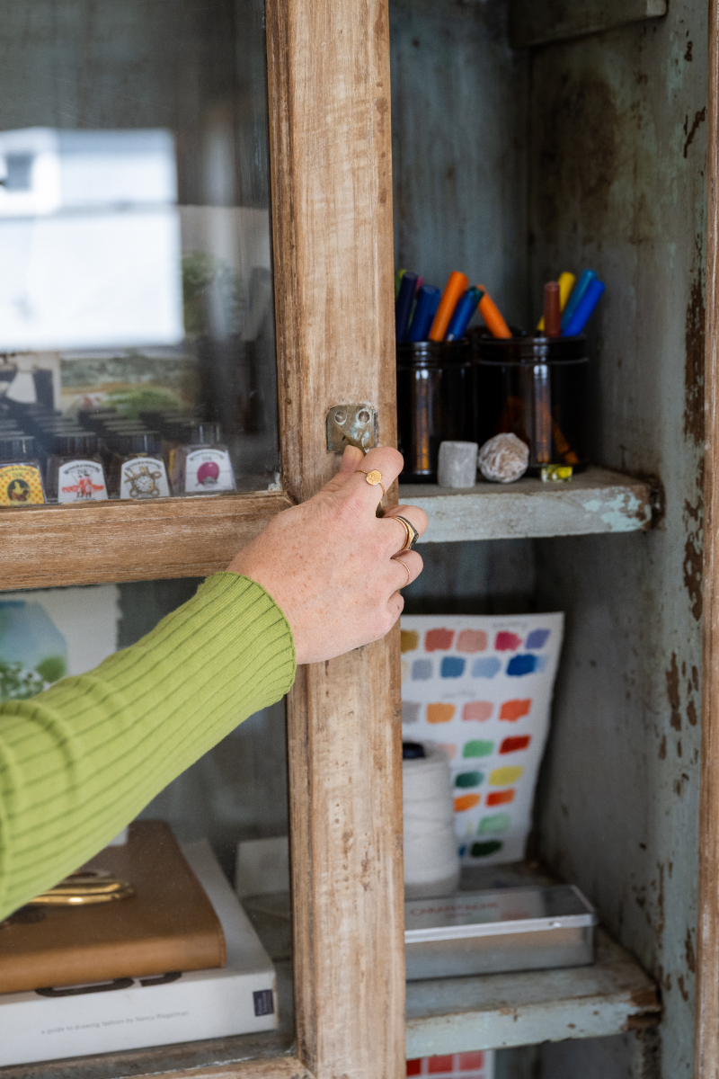 Artist opens a rustic glass-front cabinet filled with inks, markers, color swatches, and painting supplies, showing the variety of materials that can come into play when comparing cheap vs professional watercolor.
