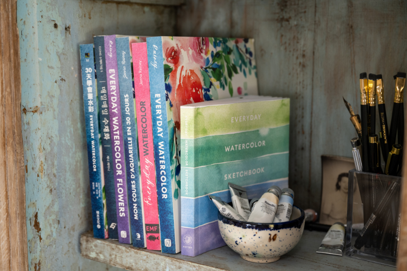 Watercolor books, a sketchbook, paint tubes, and brushes arranged on a rustic studio shelf, creating a practical collection of painting resources for artists learning about cheap vs professional watercolor.