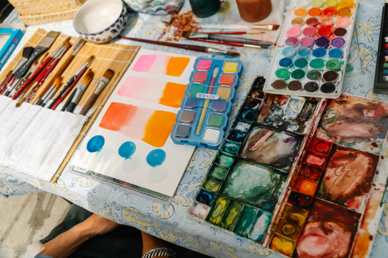 Table covered with watercolor palettes, brushes, and painted color swatches in pink, orange, blue, and green, providing a visual example of the pigments and materials used to compare cheap vs professional watercolor.