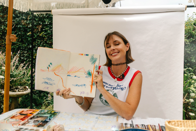 Jenna Rainey smiles while holding an open sketchbook of colorful paintings at her outdoor art table, demonstrating how watercolor mistakes are part of learning and creating art.