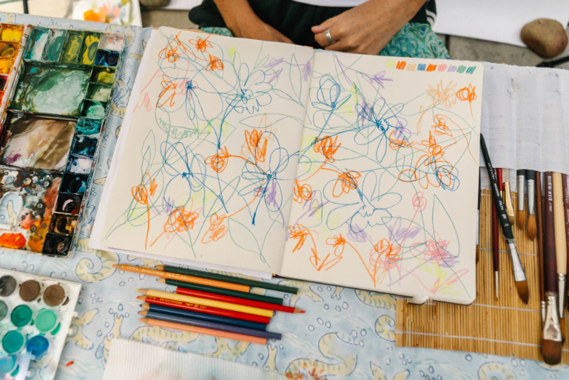 An open sketchbook filled with colorful floral doodles sits beside watercolor paints, brushes, and pencils, showing how watercolor mistakes can inspire creative experimentation.